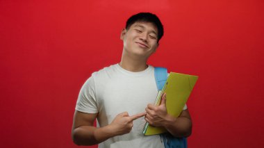 Elinde kitap tutan ve sırt çantası olan genç bir adam izole bir kırmızı arka plan duvarının üzerinde duruyor. Dostça bir gülümseme sergiliyor. Gençlik coşkusu ve coşkusu yayıyor..
