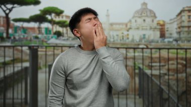 Antik Roma harabelerinin önünde esneyen genç Çinli bir adam, arka planda tarihi mimari olan bir şehir ortamında can sıkıntısını dile getiriyor..