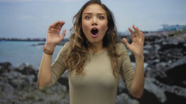 Heyecanlı yüzlü bir kadın bulutlu bir gökyüzünün altında kayalık bir sahil kenarında duruyor. Elleriyle doğanın güzelliğini ve duygularını gösteriyor..