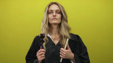 Üniformalı genç sarışın kadın yargıç, canlı sarı arka plan duvarına karşı tokmak tutuyor kapalı alan hukuku kavramına izole edilmiş