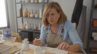 Kadın, parlak seramik stüdyosundaki ahşap masada küçük bir kil çömlek yontuyor; yaratıcı odaklanma.
