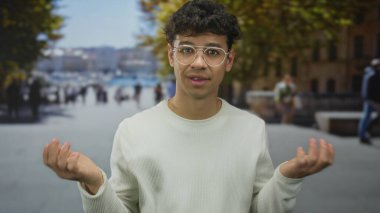 Beyaz kazaklı ve yuvarlak gözlüklü bir adam sokakta avuçlarını açık, kaşlarını kaldırmış ve başını hafifçe eğmiş olarak gösteriyor..
