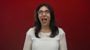 Elleri yanaklarında ve kırmızı stüdyoda ağzı açık, gözlüklü ve beyaz bluzlu bir kadın. Sürpriz..