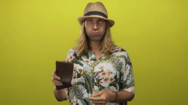 Uzun saçlı, hasır şapkalı ve çiçekli gömlekli sarışın bir adam iki eliyle bir stüdyoda cüzdanı kontrol ediyor..