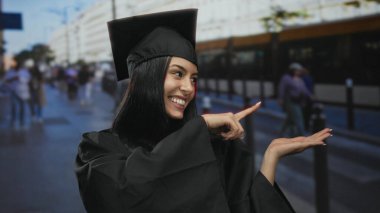 İspanyol kadın mezuniyeti başlık ve cübbe puanlarıyla kutluyor kalabalık bir şehir sokak arkaplanına karşı neşeyle, açık havada gençlik başarısının özünü yakalıyor..