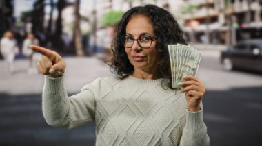 Amerikan doları tutan bir kadın, şehir dışındaki kalabalık bir sokağı işaret ederken kendine güvenen bir ifade ve orta yaş zarafetiyle..