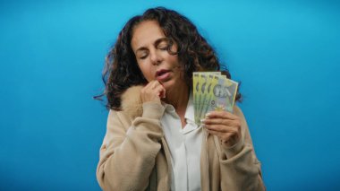 Romen banknotlarını mavi arka planda tutan orta yaşlı bir kadın yorgun görünüyor. Düşünceli bir ifadeyle finansal stresi işaret ediyor..