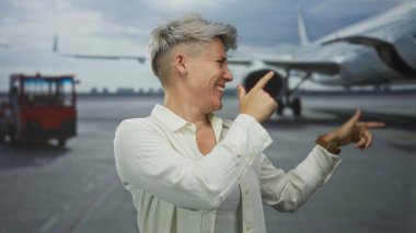 Yüzü gülen bir kadın, havaalanında pistte beyaz gömlek giyerek parmağıyla uçağı işaret ediyor; güven güçlendirme macerası.
