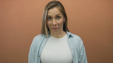 Sarı saçlı bir kadın koyu kahverengi bir arka plana karşı karmaşık bir duygu ifade ediyor. Sıradan bir kıyafetle şaşkınlık ve merak tasvir ediyor.