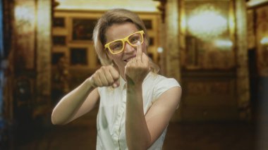 Sarı gözlüklü ve beyaz gömlekli bir kadın bina içinde orta parmağıyla kameraya doğru hareket ediyor..