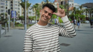 Çizgili gömlekli İspanyol adam, arka planda palmiye ağaçları ve şehir binalarıyla güneşli bir caddede rahat ve neşeli bir atmosfer sunuyor..