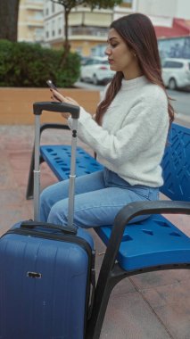 Dışarıda bir bankta bavuluyla oturan genç bir kadın, beklerken ya da şehir merkezinde seyahat ederken akıllı telefon kullanıyor..