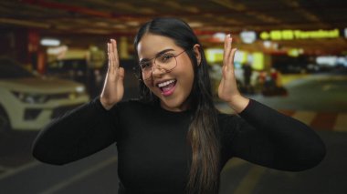 Latin asıllı genç fotoğrafçı tarafından park yerinde yakalanan kadın yüz çerçevesi. Kapalı alanda gülümsüyor. Elleri hafifçe öne çıkıyor..