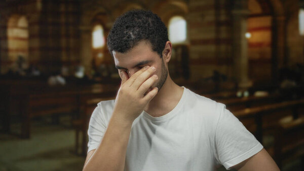 Man pinching nose with eyes closed in dimly lit stone church with arched windows; discomfort solitude.