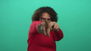 Gözlüklü ve kırmızı kazaklı bir kadın yeşil stüdyoda işaret ediyor..