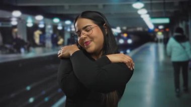 Latin bir kadın, kapalı bir tren istasyonunda neşeli bir şekilde gülümsüyor. Tren peronunun yanında kendine sarılırken sıcaklığını ve neşe saçıyor..