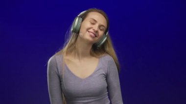 İzole edilmiş mavi bir arkaplan önünde kulaklık takan, neşeli bir ifade ve günlük kıyafetle gülümseyen ve neşeli hareketler yapan bir kadın..