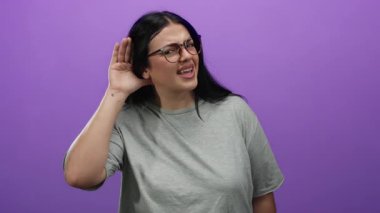 Gözlüklü genç bir kadın mor arka planda dikkatlice dinliyor izole edilmiş bir stüdyo ortamında merak ve ifade gösteriyor.