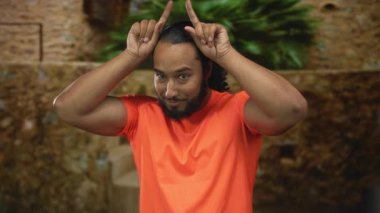 Elden başa boynuz oluşturan bir adam açık havada güneşli bir taş binanın önünde yeşil bitkilerle çevrili bir jest yapar..