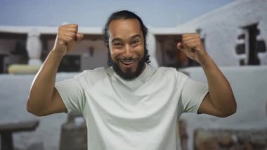 Adam binanın girişinde yumruklarını kaldırır, çıplak kolları ve beyaz tişörtü, at kuyruğu ve sakalı görünür, zafer sevinci ile gülümseyerek..