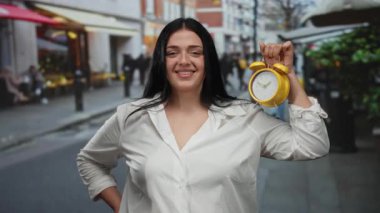 Sarı saati tutan kadın şehir caddesinde kendine güvenle gülümsüyor. Canlı arka planda açık hava gösterimi yapıyor..