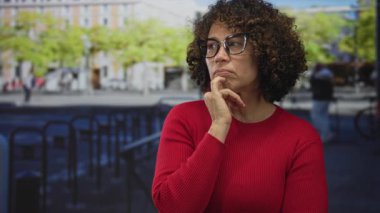 Orta yaşlı, kıvırcık saçlı, kırmızı kazak giyen İspanyol kadın, yoğun bir sokakta düşünme pozisyonunda çeneye dokunur..