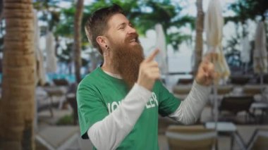 Gönüllü tişörtü giyen sakallı ve uzun kollu adam parmaklarını tatil köyü havuzuna doğru kaldırıyor. İyimserlik..