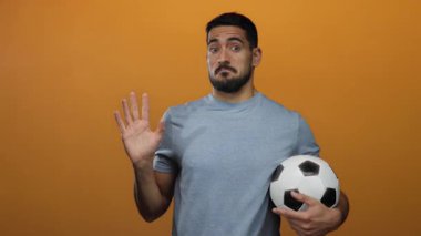 Genç adam, elinde bir futbol topuyla turuncu bir zemine doğru hareketler yapıyor. Esprili ve esprili bir sürpriz ve şaka ifadesi sergiliyor..