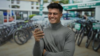 Balıkçı yaka kazaklı genç adam, bisiklet kiralama istasyonunda akıllı telefonunu kullanırken gülümsüyor. Kentin hareketli caddesinde park halindeki bisikletlerle çerçevelenmiş..