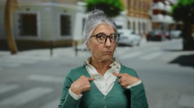 Gri saçlı ve gözlüklü yaşlı bir kadın şehir merkezinde bir sokakta durup şaşkınlığını ya da şaşkınlığını ifade ederken kendini işaret ediyor..