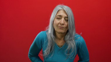 Gri saçlı, mavi kazaklı yaşlı bir kadın kırmızı arka planda rahatsız görünüyor..