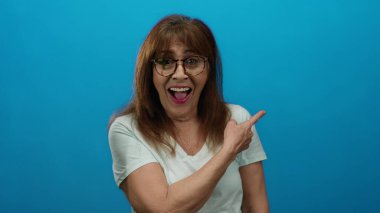 Neşeli bir ifade ve günlük bir stil sergileyen, izole bir ortamda canlı mavi bir arka plana heyecanlı bir şekilde gülümseyen ve işaret eden bir kadın..
