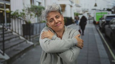 Gri kapüşonlu ve nazikçe gülümseyen kollarını göğsüne dolamış bir kadın..