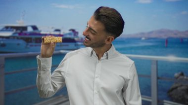 Adam güneşli iskele korkuluklarında granola çubuğunu tutuyor. Tekne gövdesi ve turkuaz deniz manzaralı. Mutluluk..
