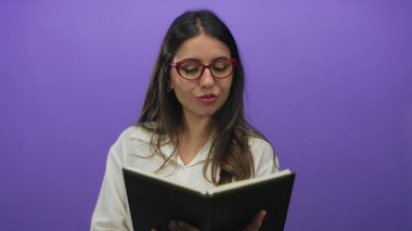 Elinde kitap tutan genç İspanyol kadın parlak mor arka planda gözlük ve beyaz bluz giyerek çekici ve entelektüel bir atmosfer yaratıyor.