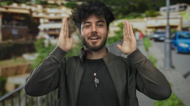 Latin asıllı genç bir adam şehir sokaklarında el hareketi yapıyor, sanatsal olarak zengin yeşillik ve arka plan mimarisi ile şehir ortamını yakalıyor..