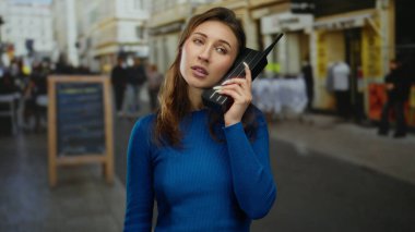 Şehir caddesinde elinde retro telefon tutan genç bir kadın, mavi giyinmiş, şehir nostaljisini canlı bir arka planda, bulanık yayalarla paylaşıyor..