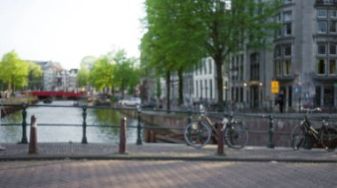 Açık bir gökyüzünün altında yeşil ağaçlı ve tarihi binalı Amsterdam Kanalı 'nın bulanık görüntüsü şehir yaşamını gözler önüne seriyor..