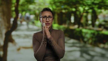 Gözlüklü kadın, güneşli bir parkta endişeyle duruyor. Etrafı yemyeşil ağaçlarla çevrili. Gün ışığında sakin bir ortamda endişe ve düşüncelerini dile getiriyor..