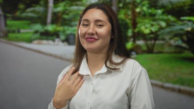 Genç İspanyol kadın, güneşli yeşil parkta hafifçe gülümserken alnının üzerinde elleriyle gözlerini koruyor..