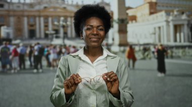 Vatikan 'ın St. Peter Meydanı' nda gülümseyen bir kadın açık gökyüzünün altında, elinde gözlüklerle, etrafı İtalyan ikonik mimarisini keşfeden turistlerle çevrili..