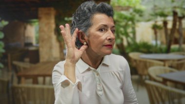 Açık hava restoranının terasında elinde ahşap mobilyalar ve doğal dekorla el kol hareketi yapan, gri saçlı, son sınıf öğrencisi dinleyen ya da kapıyı çalan, gündelik ortamda olgun duyguların altını çizen bir kadın..