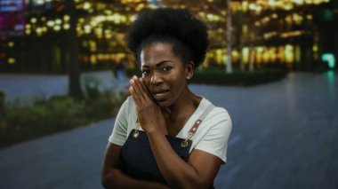 Önlüklü kadın geceleri şehir caddesinde gizli şeyler fısıldıyor. Arka planda parlak ışıklar, canlı bir şehir ortamı yaratıyor. Entrika hissini iletiyor..