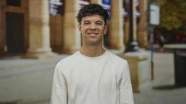 Kolları çaprazlanmış bir adam gözlük takıyor ve sokakta sütunlar inşa ederek göz kırparken gülümsüyor..