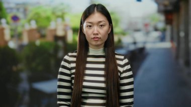 Şehir caddesinde çizgili gömlek giyen bir kadın yeşil ve bulanık arka planlı şehir ortamında düşünceli görünüyor..