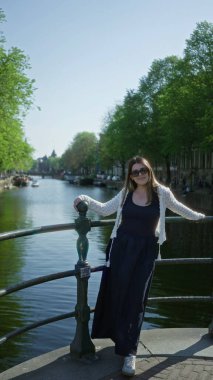Amsterdam Kanal Köprüsü 'nde dikilen kadın güneşli bir günde ağaçlarla güneşli bir günde güneşli bir elbisede açık havada gülümseyen güneş gözlükleri..