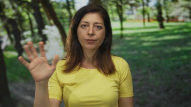 Sarı t-shirt giyen orta yaşlı İspanyol kadın güneş ışığı altında yeşil ağaçların arasında duruyor ve elini orman açıklığında sallıyor..