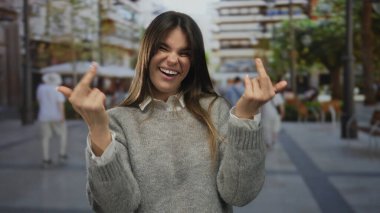 Bir kadın şehir caddesinde kaba bir jest yaparken gülümsüyor, gelişigüzel bir ortamda neşeli bir ifadeyle şehir gençlik kültürünü yakalıyor..