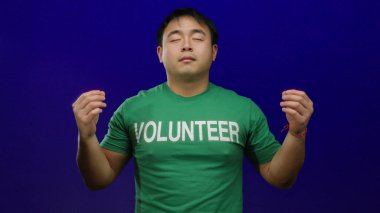 Genç bir adam yeşil bir gönüllü gömleği içinde, canlı mavi bir arka plana karşı huzur içinde meditasyon yapıyor, sakinlik ve odaklanma hissi uyandırıyor..