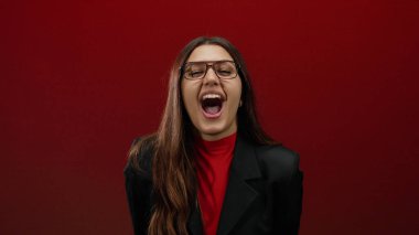 Gözlüklü, siyah ceketli, uzun saçlı, duygularını ve ifadesini yansıtan canlı kırmızı bir duvara karşı enerjik bir şekilde bağıran bir kadın..